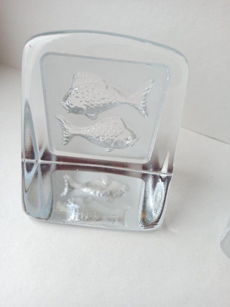 May include: A clear glass block with etched fish designs. Two fish are etched on the front, and one on the side. The glass has a frosted appearance where the fish are etched, creating a contrast with the clear glass.