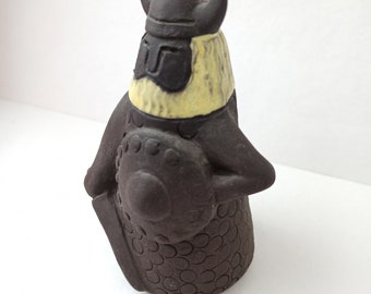 May include: A brown ceramic figurine of a Viking warrior wearing a helmet with horns and holding a shield. The figurine is detailed with a textured surface and a yellow beard.