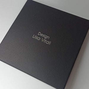 Pode incluir: Uma caixa preta com o texto "Design Lisa Vitali" em relevo em letras prateadas.