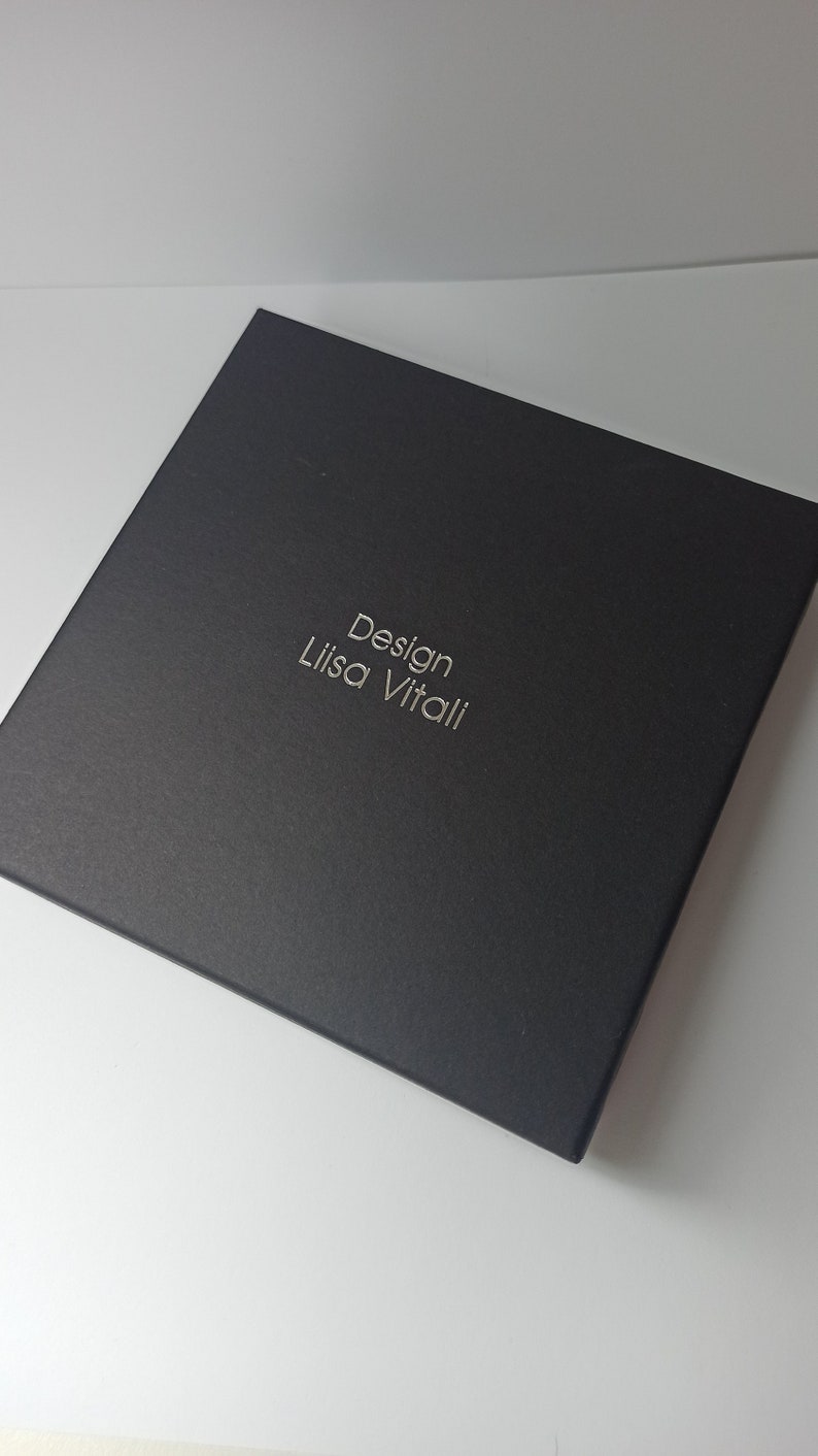 Pode incluir: Uma caixa preta com o texto "Design Lisa Vitali" em relevo em letras prateadas.