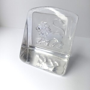 May include: A clear glass paperweight with a rectangular shape and a curved top. The paperweight features a detailed embossed image of a lion. The lion is in a standing pose, facing left. The glass is transparent, allowing light to pass through.