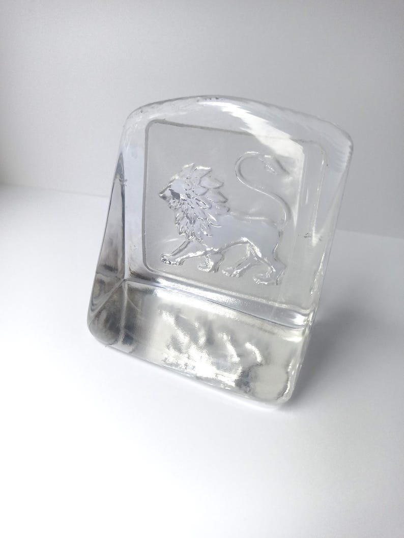 May include: A clear glass paperweight with a rectangular shape and a curved top. The paperweight features a detailed embossed image of a lion. The lion is in a standing pose, facing left. The glass is transparent, allowing light to pass through.