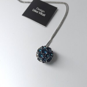 Pode incluir: Um colar de corrente de prata com um pingente redondo feito de pequenas flores de metal azul escuro. O pingente tem um di&acirc;metro de aproximadamente 2,5 cm. O colar est&aacute; sobre um fundo branco com um cart&atilde;o preto que diz "Design Lisa Vitali".