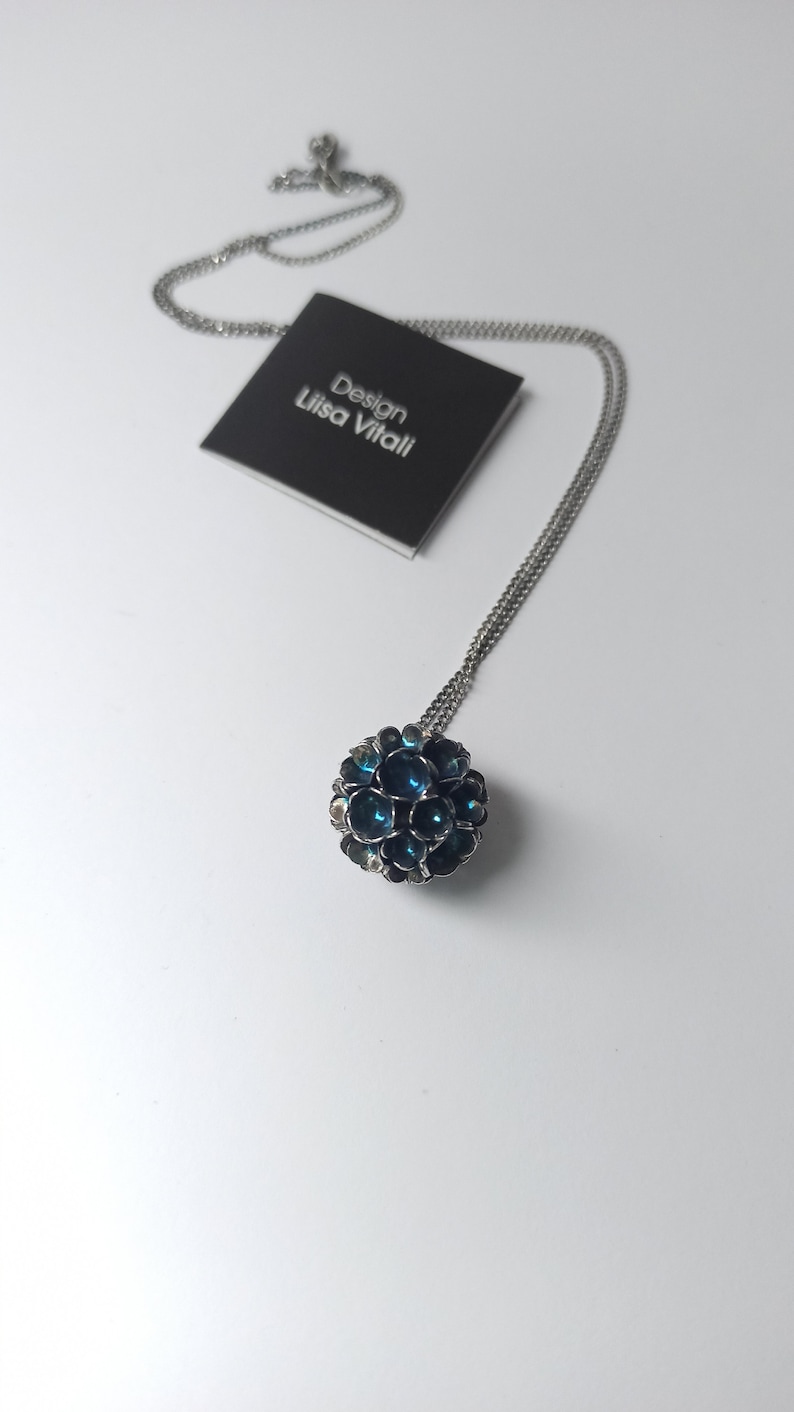 Pode incluir: Um colar de corrente de prata com um pingente redondo feito de pequenas flores de metal azul escuro. O pingente tem um di&acirc;metro de aproximadamente 2,5 cm. O colar est&aacute; sobre um fundo branco com um cart&atilde;o preto que diz "Design Lisa Vitali".