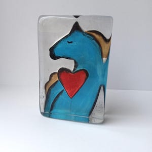Peut inclure: Un bloc de verre transparent avec un dessin peint d'un cheval bleu avec un cœur rouge. Le cheval a une crinière dorée et des contours noirs. Le verre est rectangulaire et se tient debout.