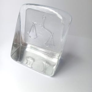 May include: A clear, rectangular glass paperweight with a slightly curved top. The paperweight features an embossed image of scales, a symbol of justice. The glass is transparent, allowing light to pass through, and sits on a white surface.