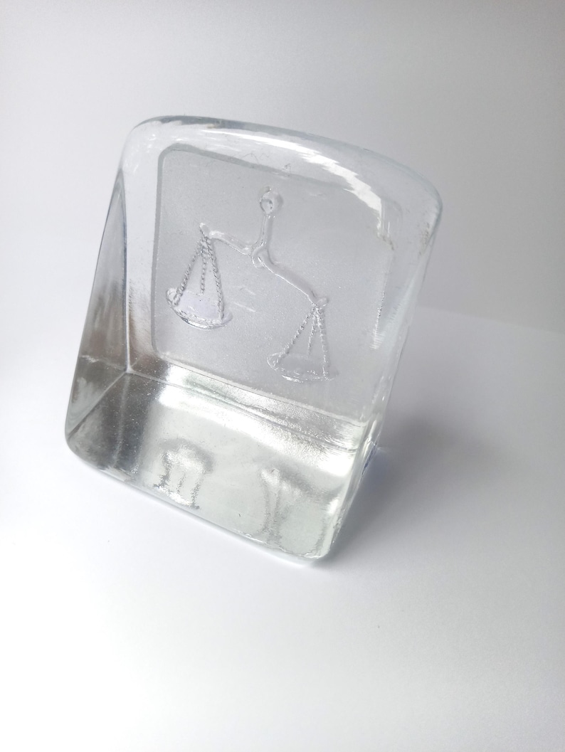 May include: A clear, rectangular glass paperweight with a slightly curved top. The paperweight features an embossed image of scales, a symbol of justice. The glass is transparent, allowing light to pass through, and sits on a white surface.