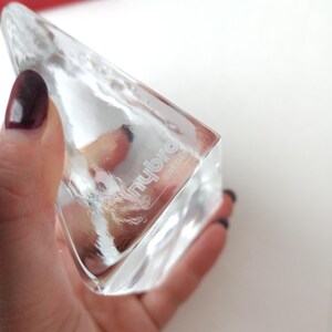 May include: A clear, triangular glass object with the text "nybro" and "sweden" etched on the side. The object is held in a hand with dark nail polish. The glass has a textured surface and is partially transparent.