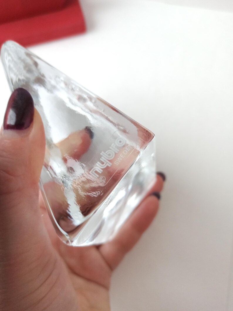 May include: A clear, triangular glass object with the text "nybro" and "sweden" etched on the side. The object is held in a hand with dark nail polish. The glass has a textured surface and is partially transparent.