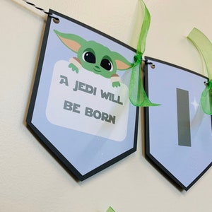 May include: A blue and black banner with a green character holding a sign that says "A Jedi Will Be Born". The banner is decorated with green ribbon.
