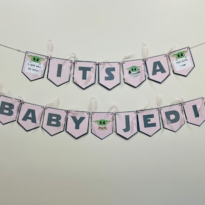 May include: Pink and gray banner with the words "It's a Baby Jedi" and images of a green alien character. The banner has pink ribbon and a string to hang it.