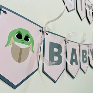 May include: Pink and gray baby shower banner with the words "It's a Baby" and a green character with big eyes.