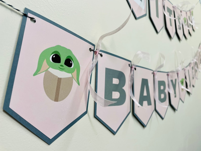 May include: Pink and gray baby shower banner with the words "It's a Baby" and a green character with big eyes.