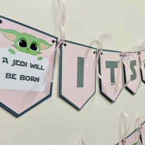 May include: Pink and gray banner with the text "It's a Jedi Will Be Born" and a cartoon image of a green baby Yoda. The banner is strung with pink ribbon.
