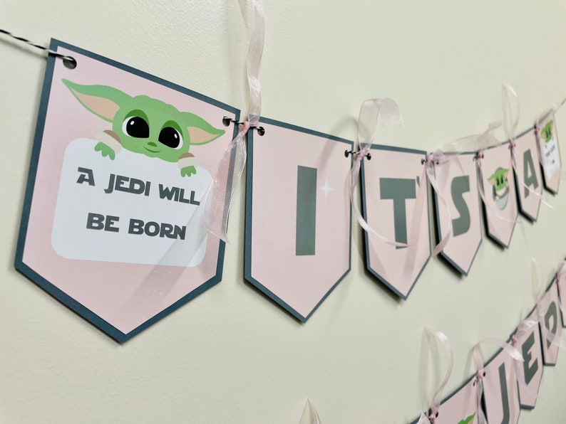 May include: Pink and gray banner with the text "It's a Jedi Will Be Born" and a cartoon image of a green baby Yoda. The banner is strung with pink ribbon.