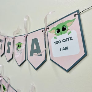 May include: Pink and gray banner with the words "It's a" and a cartoon character holding a sign that says "Too Cute I Am".