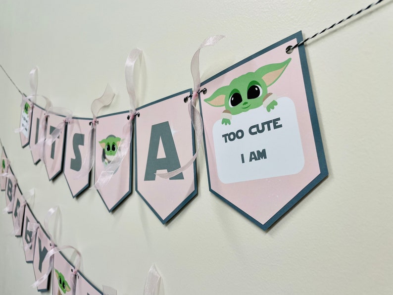 May include: Pink and gray banner with the words "It's a" and a cartoon character holding a sign that says "Too Cute I Am".