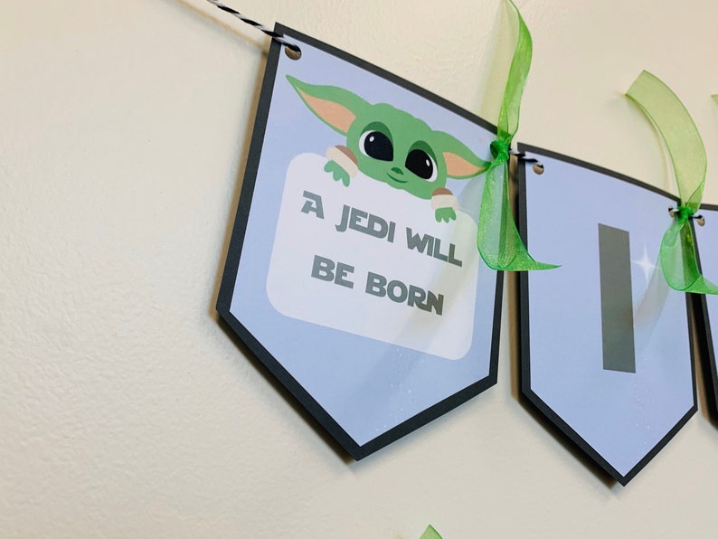 May include: A blue and black banner with a green character holding a sign that says "A Jedi Will Be Born". The banner is decorated with green ribbon.