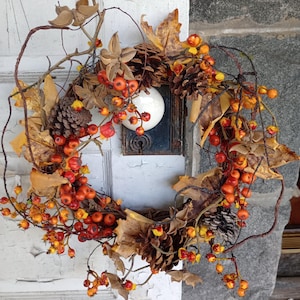 May include: A fall-themed wreath with a mix of natural elements. It features brown pinecones, dried leaves, and vibrant orange berries. The wreath is set against a white door with peeling paint and a stone wall.