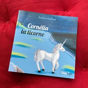 Peut inclure: Un livre pour enfants bleu et blanc intitulé "Cornélia la licorne" de LalexAndrea. Le livre présente une illustration d'une licorne blanche avec une corne arc-en-ciel sur un fond bleu avec des étoiles.