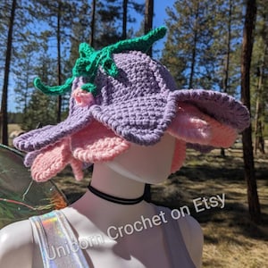 May include: A handmade crochet flower hat in shades of lavender, pink, and green. The hat features a wide brim and is displayed on a mannequin head. The text "Unicorn Crochet on Etsy" is visible.