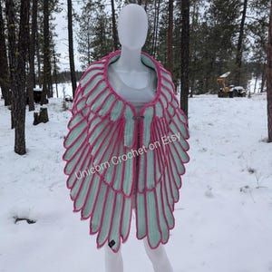 Könnte beinhalten: Ein gehäkeltes Tuch mit einem Flügel-ähnlichen Design in Rosa- und Hellgrüntönen. Das Tuch wird auf einer weißen Schaufensterpuppe vor einem schneebedeckten Hintergrund präsentiert. Der Text "Unicorn Crochet on Etsy" ist auf dem Tuch sichtbar.