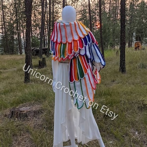 Könnte beinhalten: Ein Regenbogen-Häkel-Schal mit einem gewellten Rand. Der Schal ist über eine weiße Schaufensterpuppe in einer Waldlandschaft drapiert.