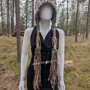 May include: A beige crochet hat with long, braided strands. The hat features a textured design and is adorned with decorative elements. The braids are a mix of brown, beige, and blue fibers. The text "Unicorn Crochet on Etsy" is visible on the dress.