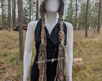 Puede incluir: Un gorro de ganchillo beige con largas trenzas. El gorro presenta un diseño texturizado y está adornado con elementos decorativos. Las trenzas son una mezcla de fibras marrones, beiges y azules. El texto "Unicorn Crochet on Etsy" es visible en el vestido.