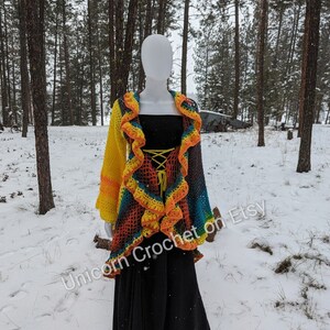 May include: A colorful crochet cardigan with a ruffled trim. The cardigan is made in a rainbow of colors, including yellow, orange, red, blue, and green. The cardigan is shown on a mannequin in a snowy forest setting. The text "Unicorn Crochet on Etsy" is visible in the lower right corner of the image.