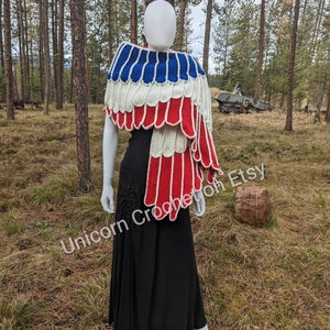 Könnte beinhalten: Ein gehäkelter Schal in Rot, Weiß und Blau, der an die amerikanische Flagge erinnert, über einem weißen Mannequin in einer Waldlandschaft drapiert. Der Schal ist mit einem gewellten Rand gearbeitet und hat ein einzigartiges Design.