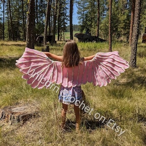 Könnte beinhalten: Ein Kind trägt ein pinkfarbenes und weißes gehäkeltes Einhornflügelkostüm in einer Waldlandschaft. Die Flügel sind groß und gefiedert, und das Kind hält sie seitlich ausgestreckt. Der Text "Unicorn Crochet on Etsy" ist in der unteren rechten Ecke des Bildes sichtbar.