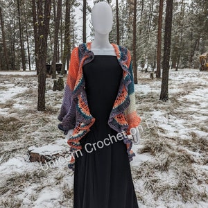 May include: A crocheted shawl with a ruffled edge in shades of purple, teal, orange, and white. The shawl is draped over a black dress form in a snowy forest setting.