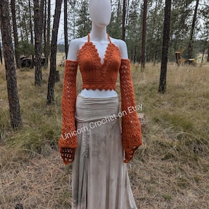 May include: A burnt orange crochet top with long bell sleeves and a halter neckline. The top is worn over a long, flowing, light beige skirt. The model is standing in a forest setting.