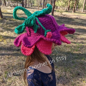 May include: A crocheted flower hat in shades of pink, purple, and green. The hat has a large flower design with a stem and leaves. The hat is worn by a person with long brown hair. The text "Unicorn Crochet on Etsy" is visible in the image.
