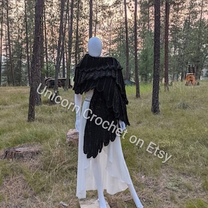 Könnte beinhalten: Ein schwarzer Häkel-Schal mit einem gekräuselten Rand, drapiert über eine weiße Schaufensterpuppe. Der Schal wird in einer Waldlandschaft präsentiert.