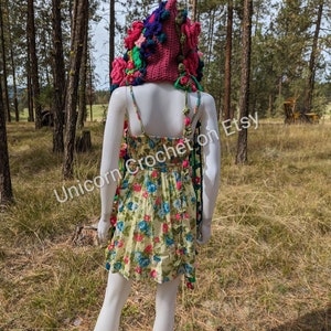 May include: A pink crocheted hooded scarf with colourful flowers and pom-poms. The scarf is displayed on a white mannequin in a woodland setting. The text "Unicorn Crochet on Etsy" is visible in the image.