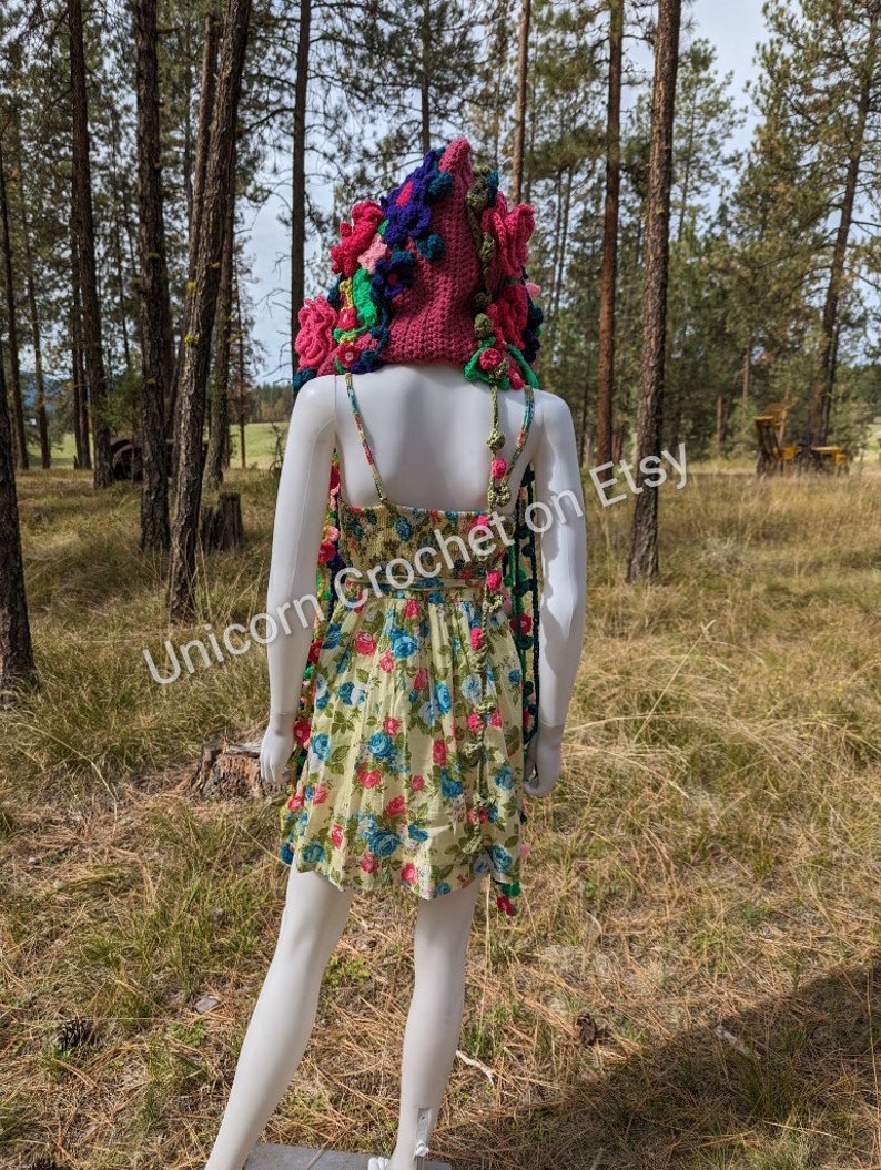 May include: A pink crocheted hooded scarf with colourful flowers and pom-poms. The scarf is displayed on a white mannequin in a woodland setting. The text "Unicorn Crochet on Etsy" is visible in the image.