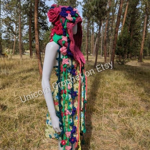 May include: A pink crocheted hooded scarf with colourful flowers and vines. The scarf is worn by a mannequin in a floral dress.  "Unicorn Crochet on Etsy" is written on the scarf.