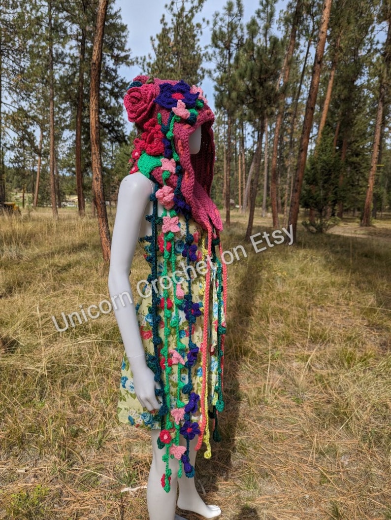 May include: A pink crocheted hooded scarf with colourful flowers and vines. The scarf is worn by a mannequin in a floral dress.  "Unicorn Crochet on Etsy" is written on the scarf.