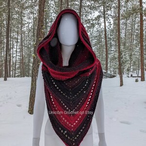 May include: A red, black, and grey hooded crochet shawl with a lacy pattern. The shawl is draped over a white mannequin in a snowy forest setting. The text "Unicorn Crochet on Etsy" is visible at the bottom of the image.