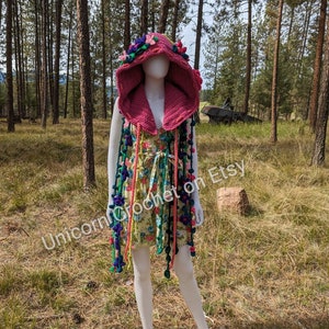 May include: A pink crocheted hooded vest with colourful floral embellishments and long fringe. The vest is worn on a white mannequin in a woodland setting.