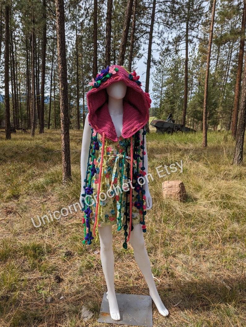 May include: A pink crocheted hooded vest with colourful floral embellishments and long fringe. The vest is worn on a white mannequin in a woodland setting.