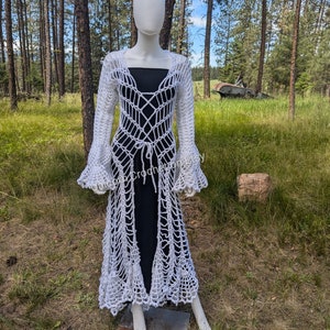May include: White crochet long-sleeved cardigan with a tie at the waist and bell sleeves. The cardigan is shown on a white mannequin against a background of trees and grass.
