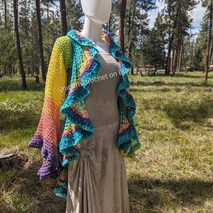 May include: A rainbow crochet shawl with a ruffled edge. The shawl is draped over a white mannequin and is displayed against a backdrop of green trees.