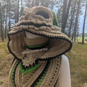 May include: A handmade crochet hood and scarf set in tan, black, and green. The hood features a face-like design with black trim and a green accent. The scarf has a matching pattern. The text "Unicorn Crochet on Etsy" is visible.