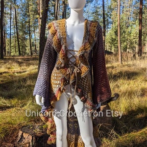 May include: A long, crocheted cardigan with a high neckline, a lace-up front, and a ruffled hem. The cardigan is made in a variety of colors, including purple, brown, and yellow. The cardigan is shown on a white mannequin in a forest setting.
