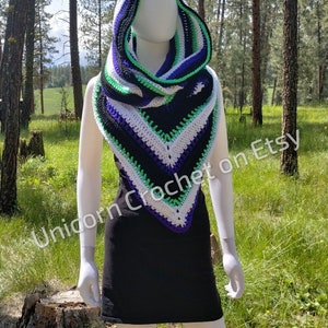 May include: A hooded scarf in a triangular shape with stripes of black, white, purple, and green yarn. The scarf is crocheted and is being modeled by a white mannequin in a black skirt. The mannequin is standing in a forest setting.