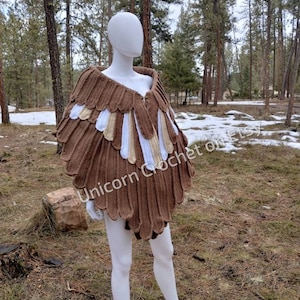 Könnte beinhalten: Ein braunes Häkeltuch mit weißen und beigefarbenen Akzenten. Das Tuch ist in Form eines Vogelflügels geformt und hat einen gewellten Rand. Das Tuch wird auf einem weißen Mannequin in einer Waldlandschaft präsentiert.