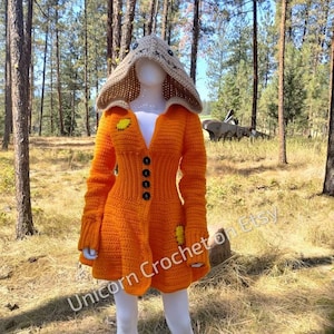 May include: Orange crochet cardigan with a brown crocheted hood featuring a stitched face. The cardigan has a button closure and a flared skirt. The text "Unicorn Crochet on Etsy" is visible in the bottom left corner of the image.